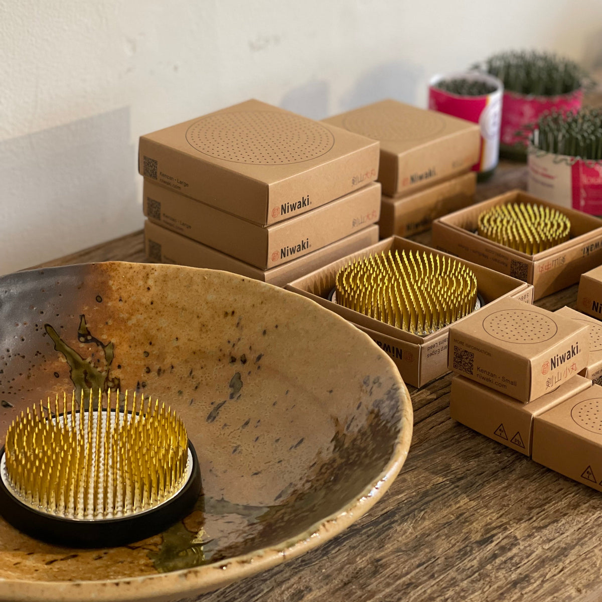 A set of Niwaki kenzan flower frog pin holders, displayed alongside their brown kraft packaging boxes on a rustic wooden table. A large, textured Wabi sabi brown ceramic bowl made in Japan is in the foreground.