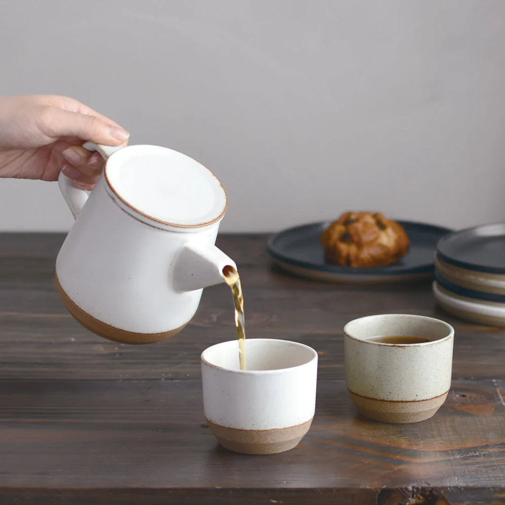 Person pouring tea from a white ceramic teapot into two cups on a wooden table.