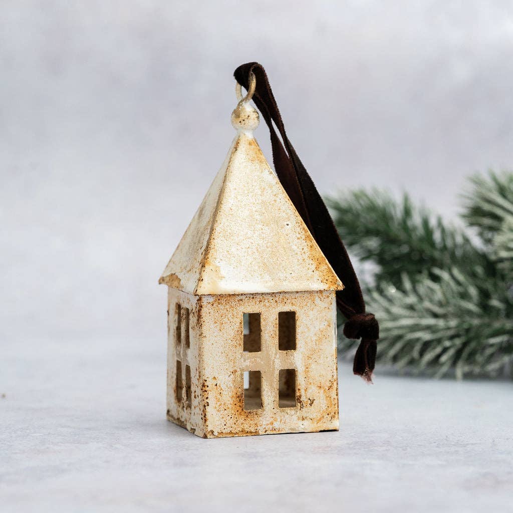 A small decorative house-shaped ornament with a distressed cream and brown finish and a dark brown hanging ribbon, photographed against a soft light gray background with blurred greenery in the back.