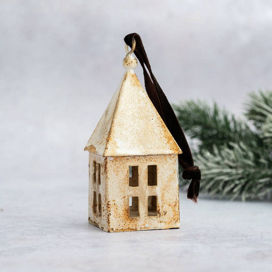 A small decorative house-shaped ornament with a distressed cream and brown finish and a dark brown hanging ribbon, photographed against a soft light gray background with blurred greenery in the back.