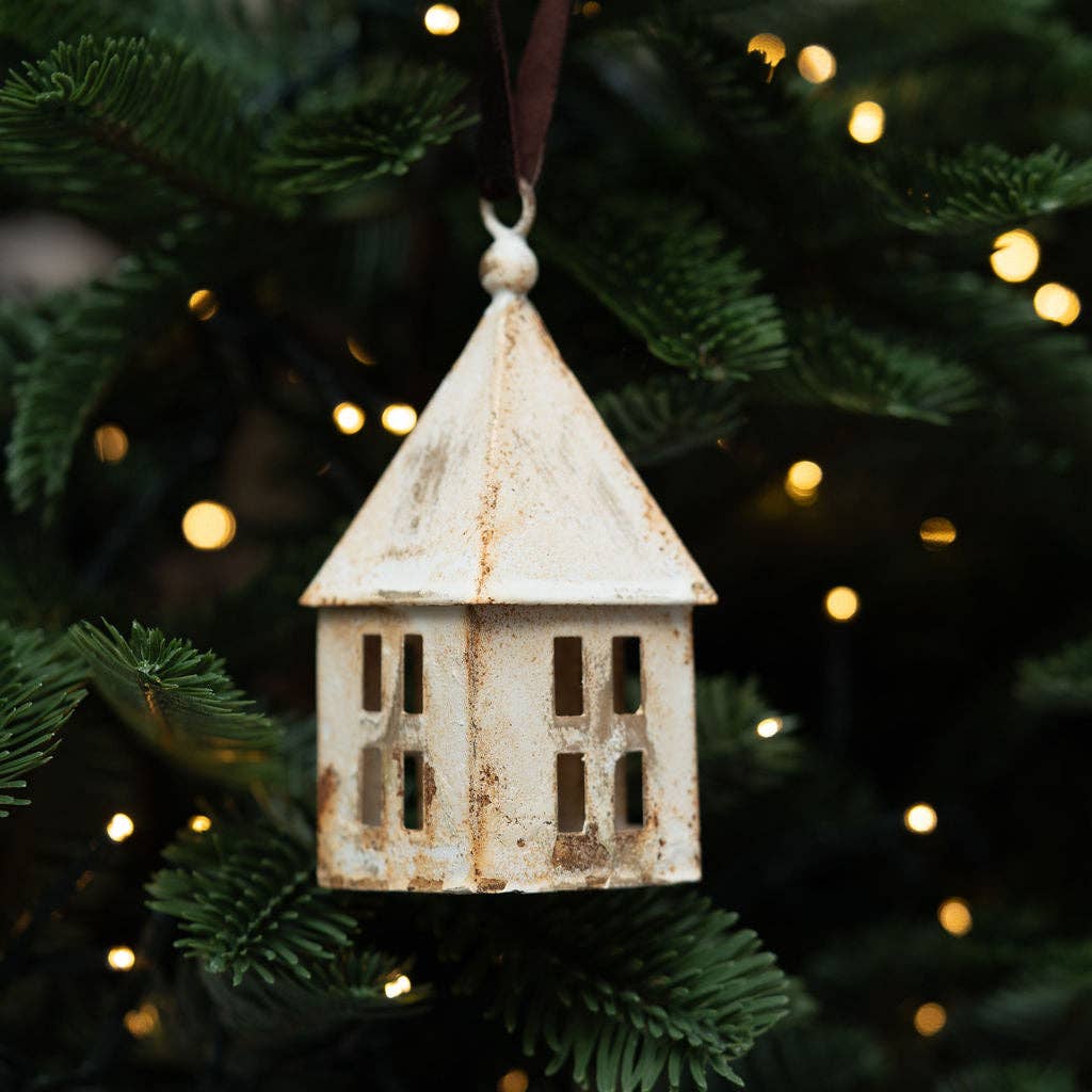 A rustic white Christmas ornament hanging from a dark brown ribbon on a green Christmas tree with warm glowing lights in the background.