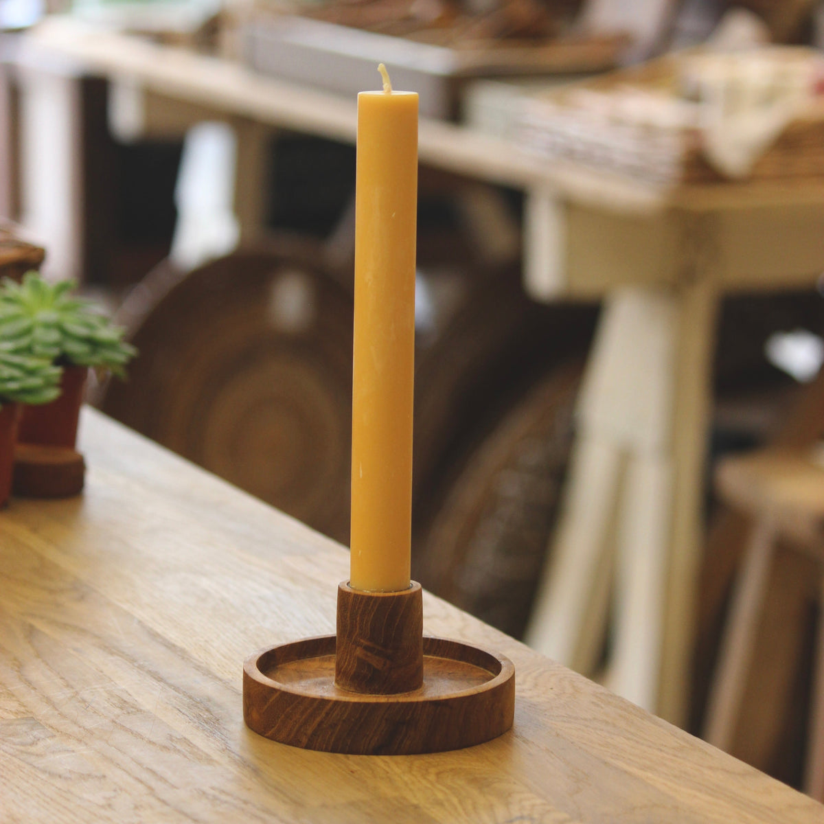 A yellow plant-based dinner candle standing upright in a Luna round teak candle holder on a wooden table, with a softly blurred workshop or interior background.