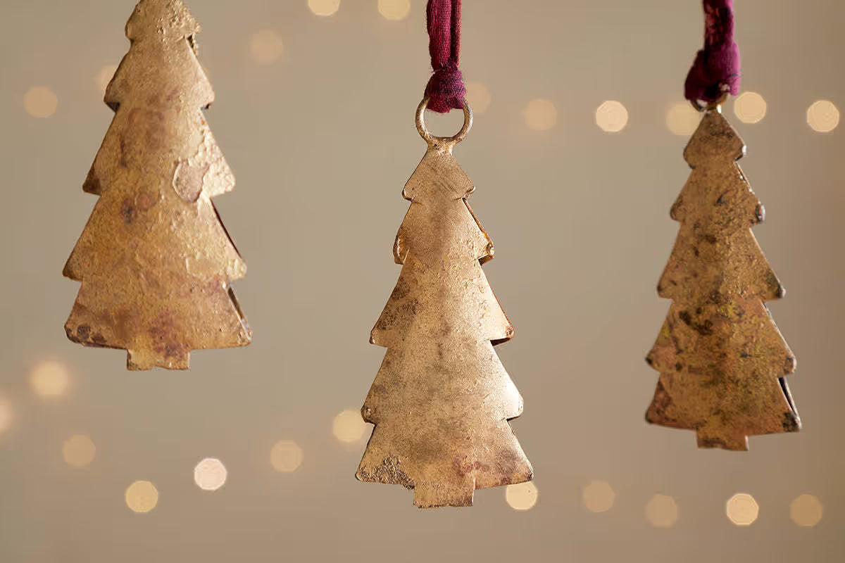 Three gold Christmas tree-shaped ornaments hanging against a soft, blurred background with lights. Four gold Christmas tree ornaments nkuku Mishti Tree Baubles 