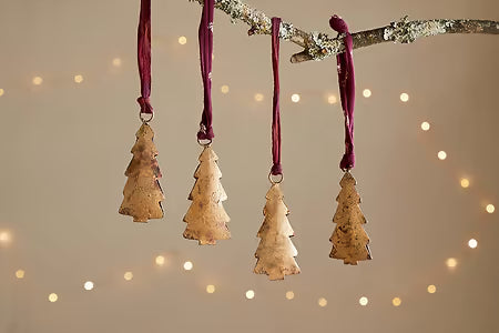 Four gold Christmas tree ornaments nkuku Mishti Tree Baubles hanging from a branch against a warm, blurred light background.