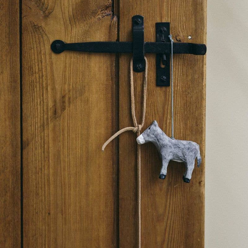 A small grey papier mache donkey made in India, hanging from a rope attached to a black metal latch on a rustic wooden door.