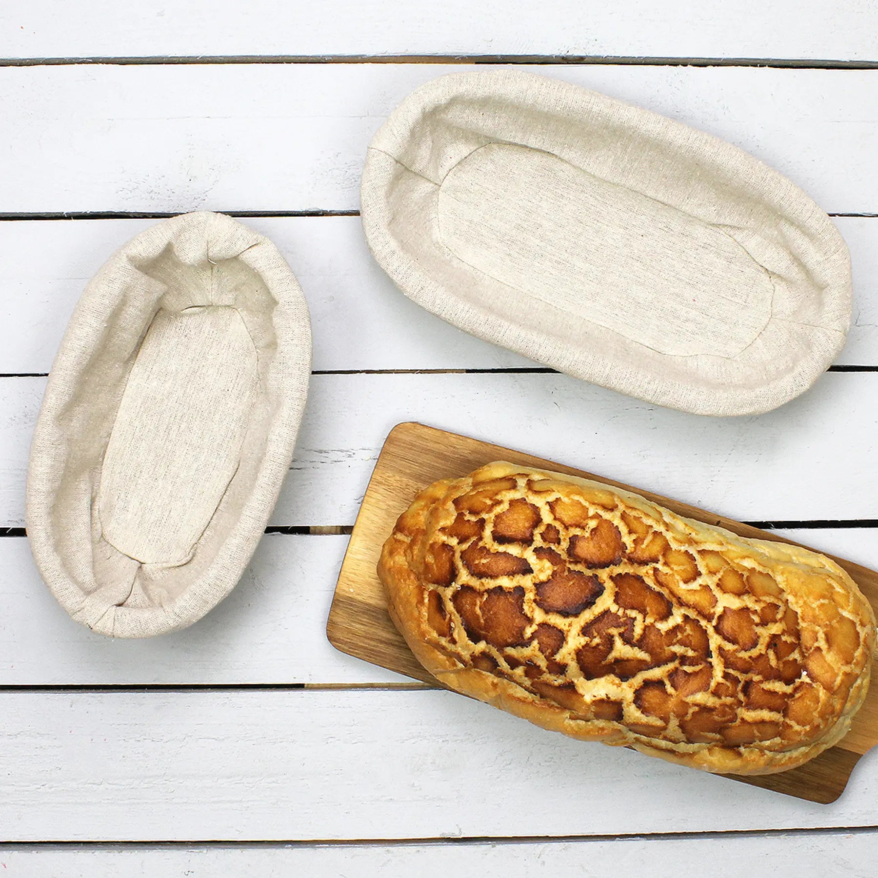 Two bread proofing baskets on a wooden surface with a loaf of bread on a cutting board.