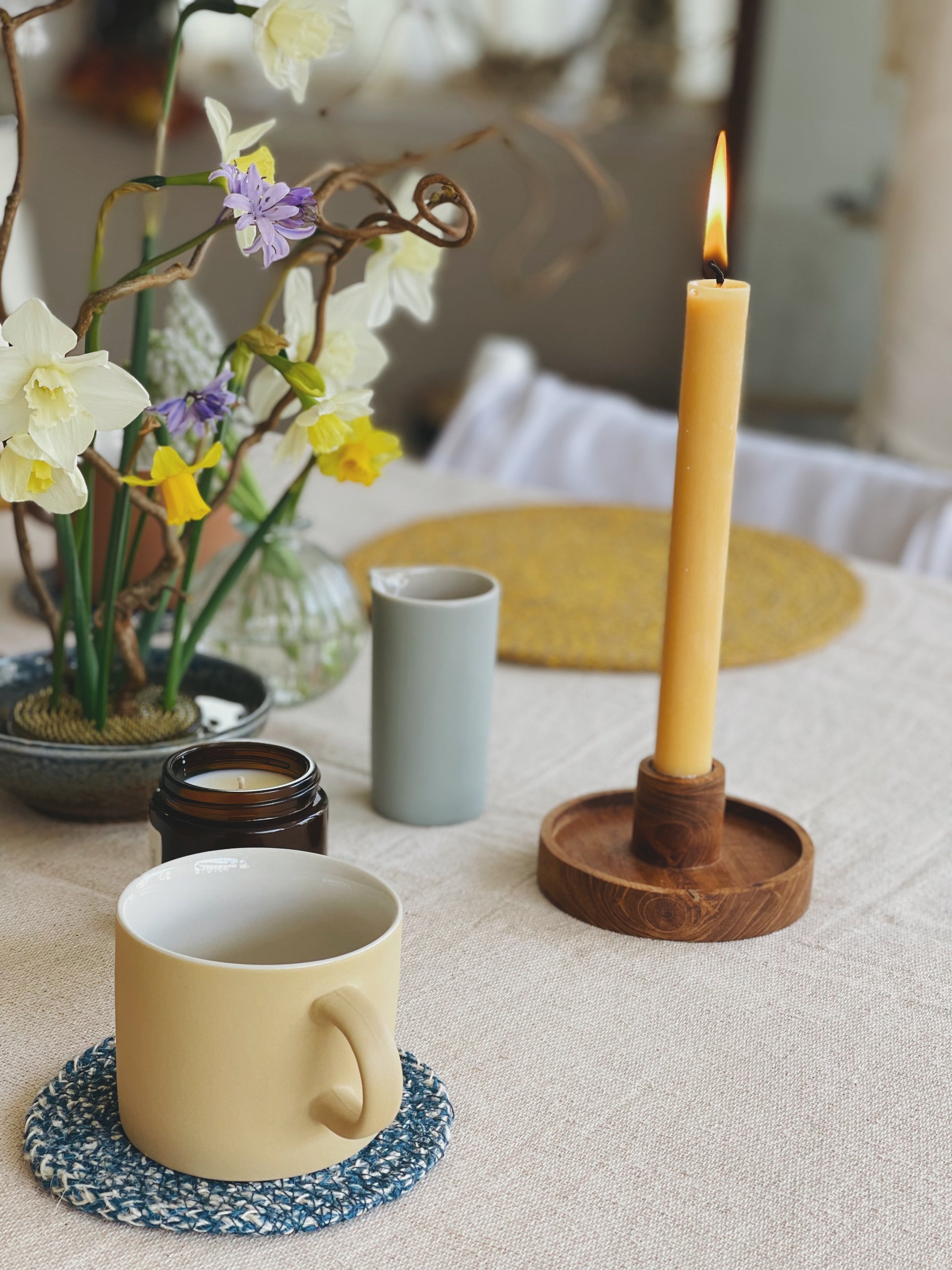 Candle in a wooden holder on a table with a mug, flowers, and a teacup.