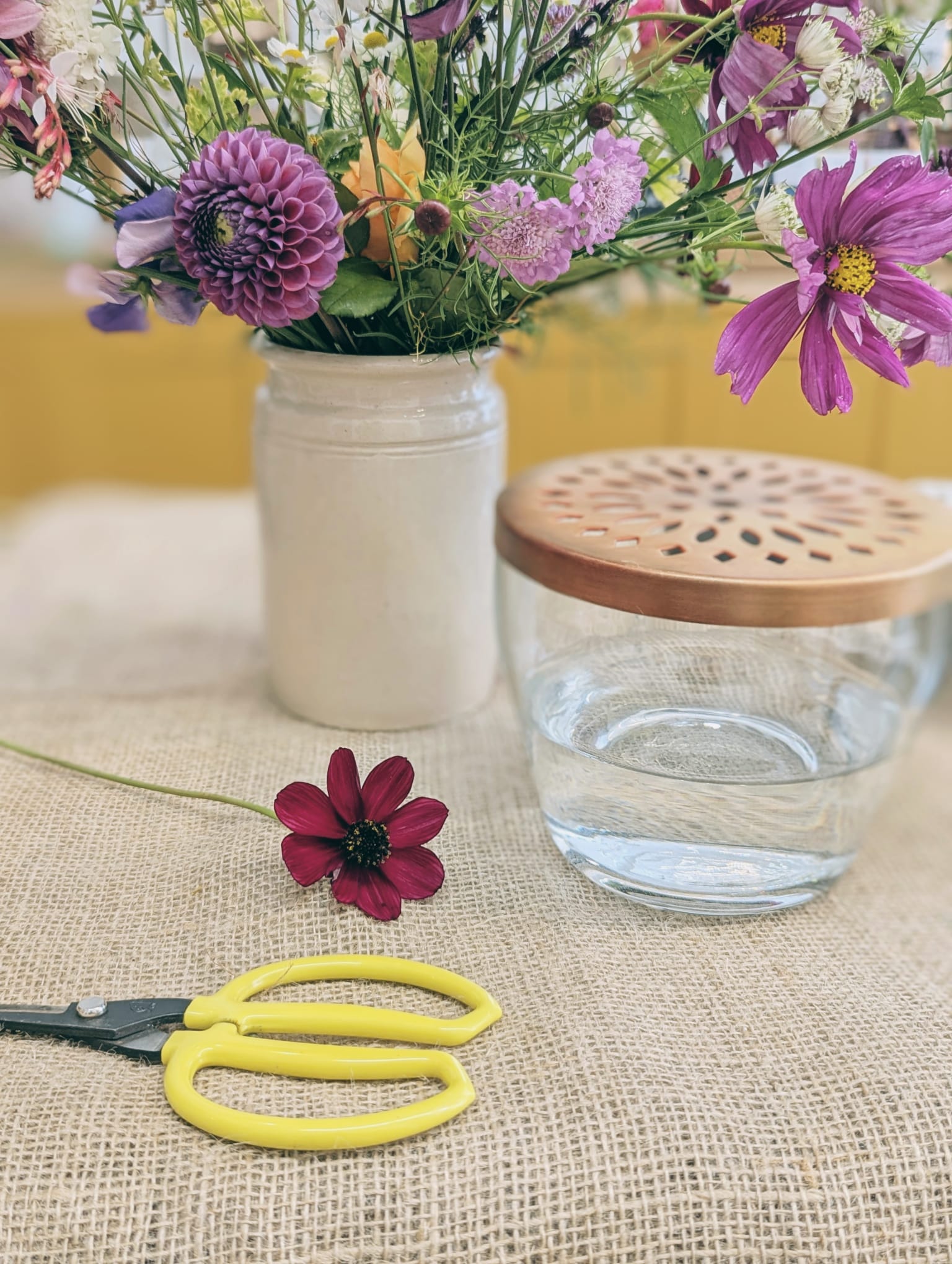 Glass jar with brass lid, scissors, and a small flower on a textured surface with a blurred floral background.