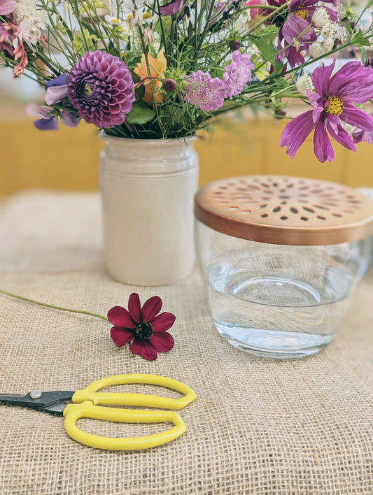 Glass jar with brass lid, scissors, and a small flower on a textured surface with a blurred floral background.