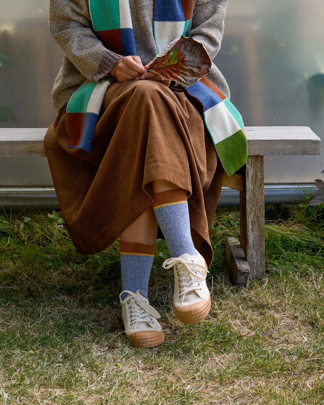 Person sitting on a bench wearing a colorful scarf, brown skirt, blue Rove socks, and white sneakers.
