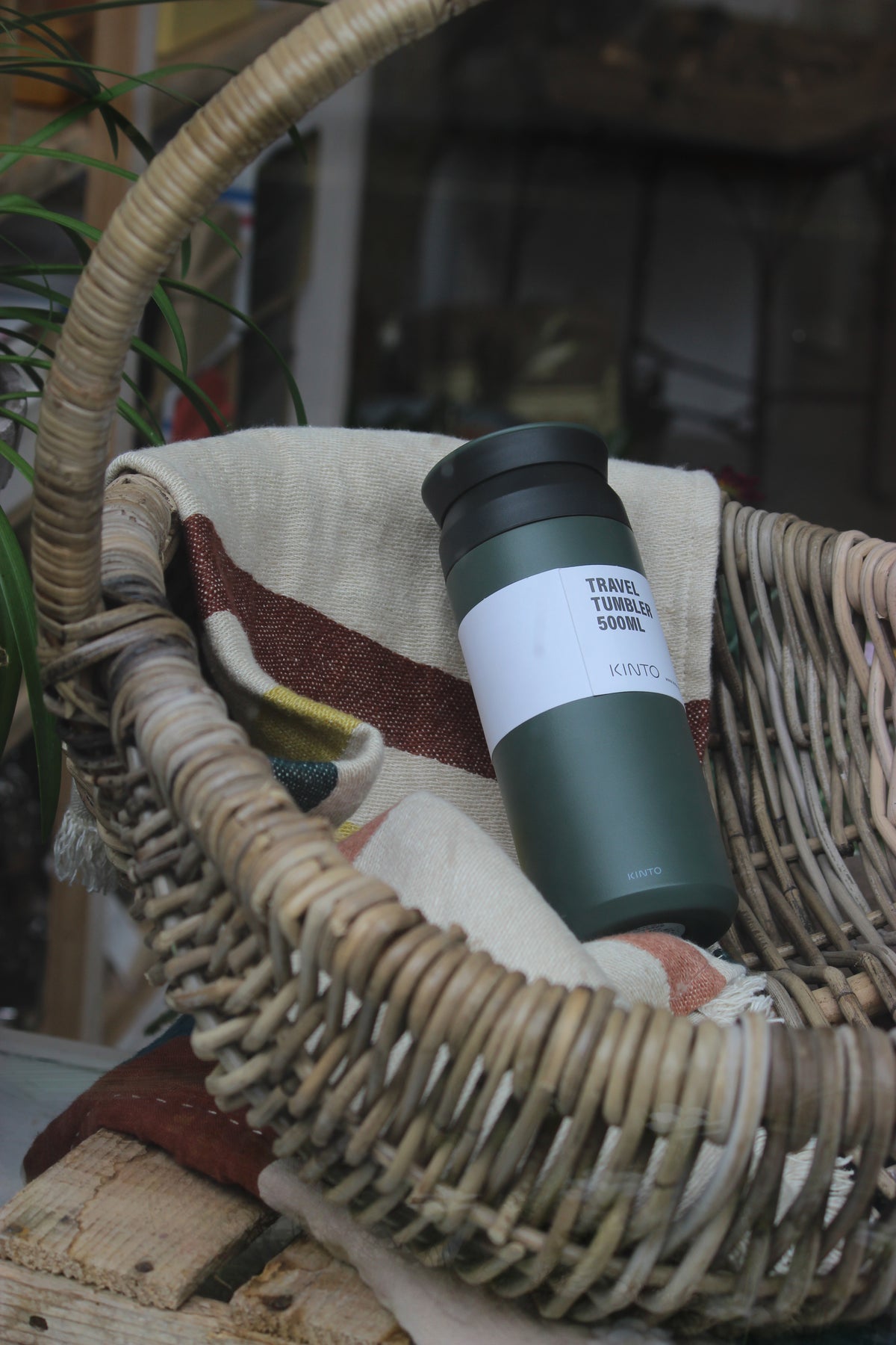 A dark green insulated travel tumbler with a white label reading 'TRAVEL TUMBLER 500ML' and 'KINTO', resting inside a woven wicker basket on a beige cloth.