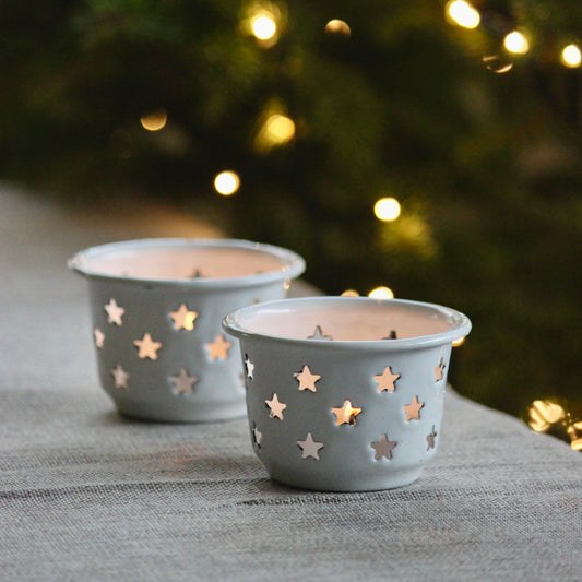 Two cream enamel star-shaped candle holders with cutout star patterns, shown lit on a grey fabric surface with a blurred Christmas tree and warm string lights in the background.