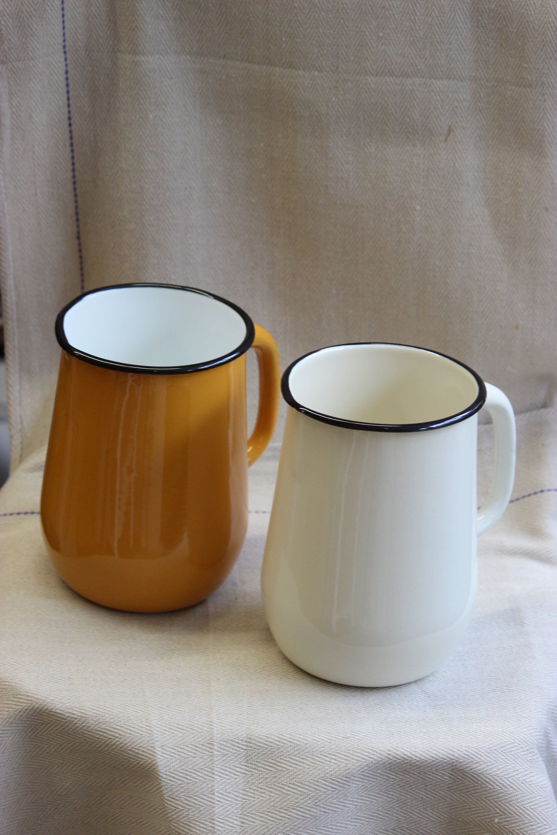 Two enamel jugs, one white and one mustard, with black rims, on a textured fabric background.