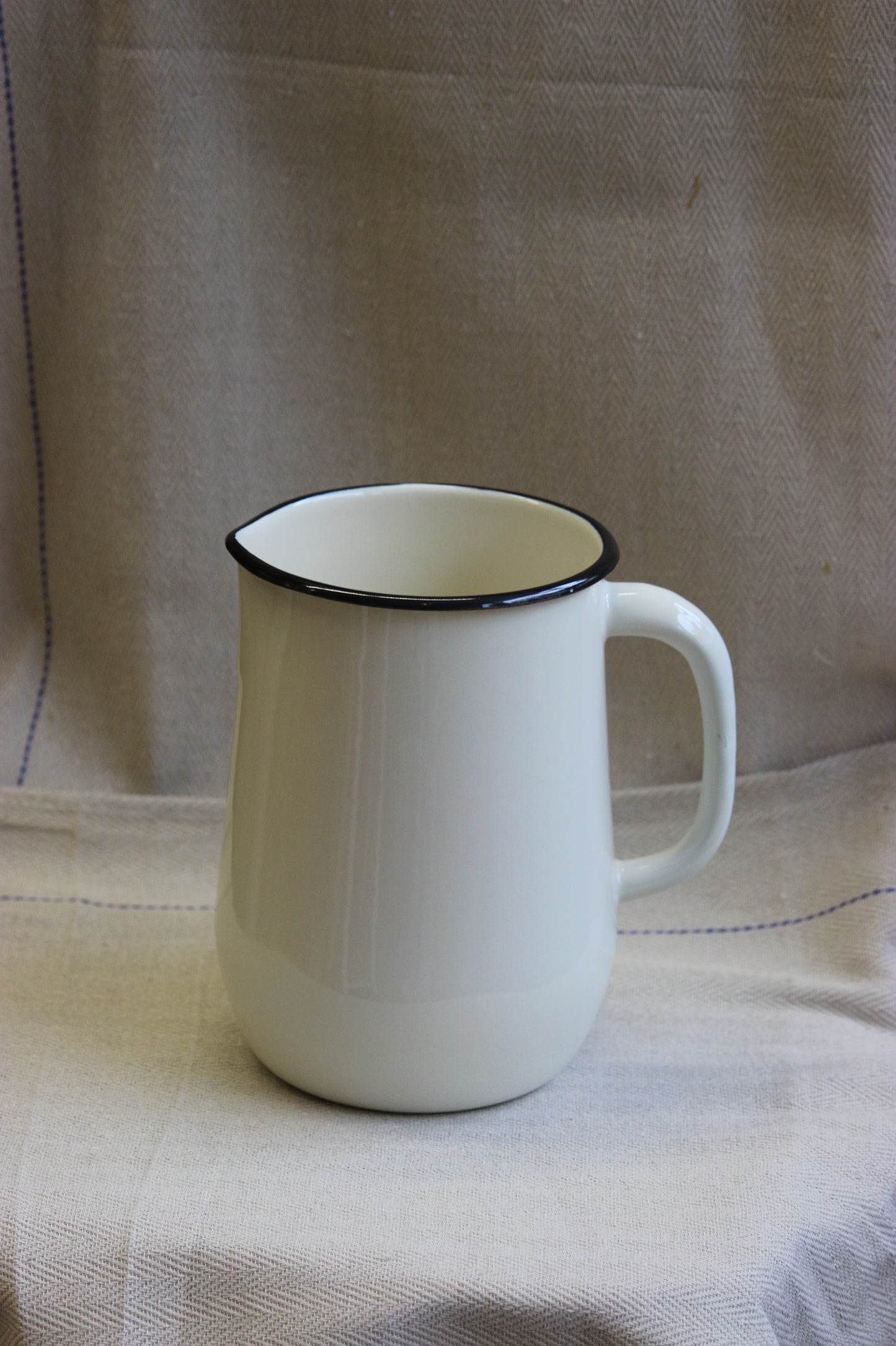 White cream enamelware jug made in Ukraine with a black rim on a beige fabric background