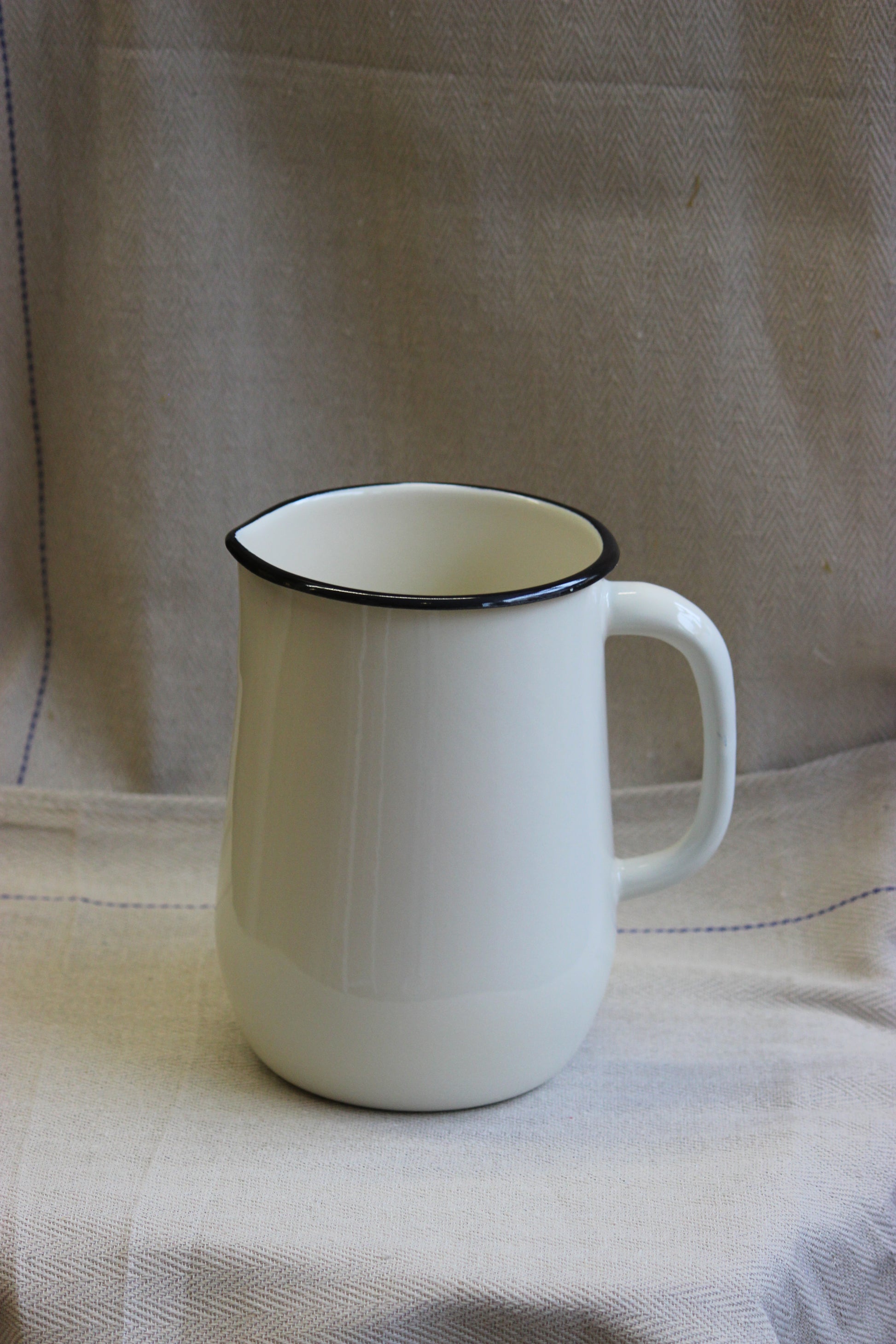 White cream enamelware jug made in Ukraine with a black rim on a beige fabric background