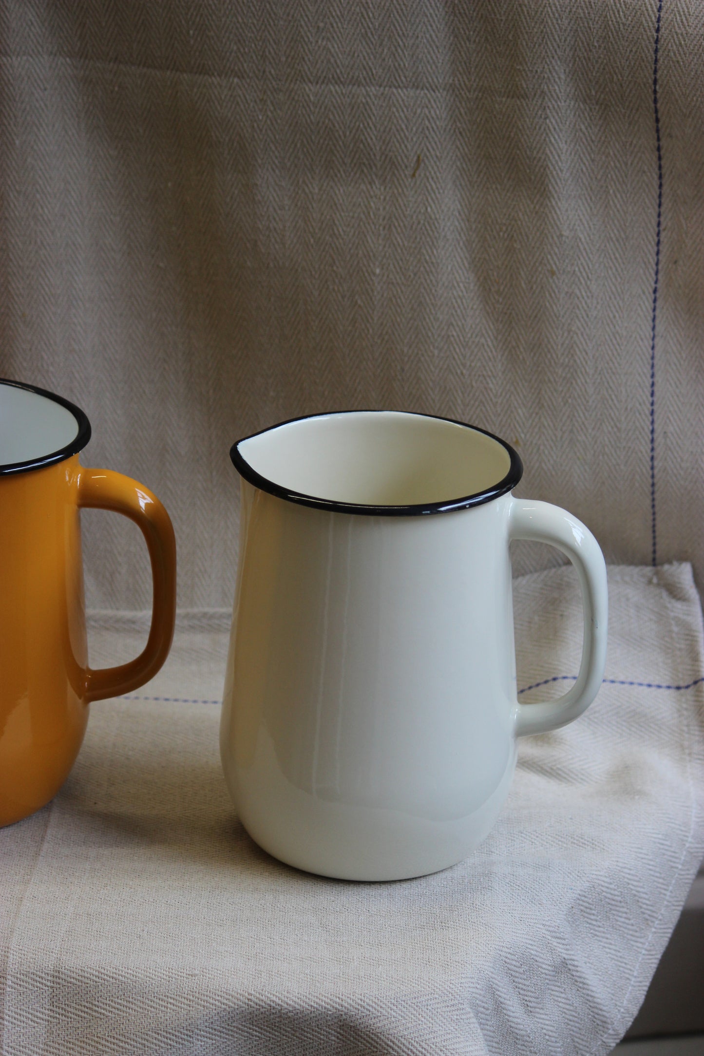 Two enamel jugs, one white and one yellow, on a textured fabric background.