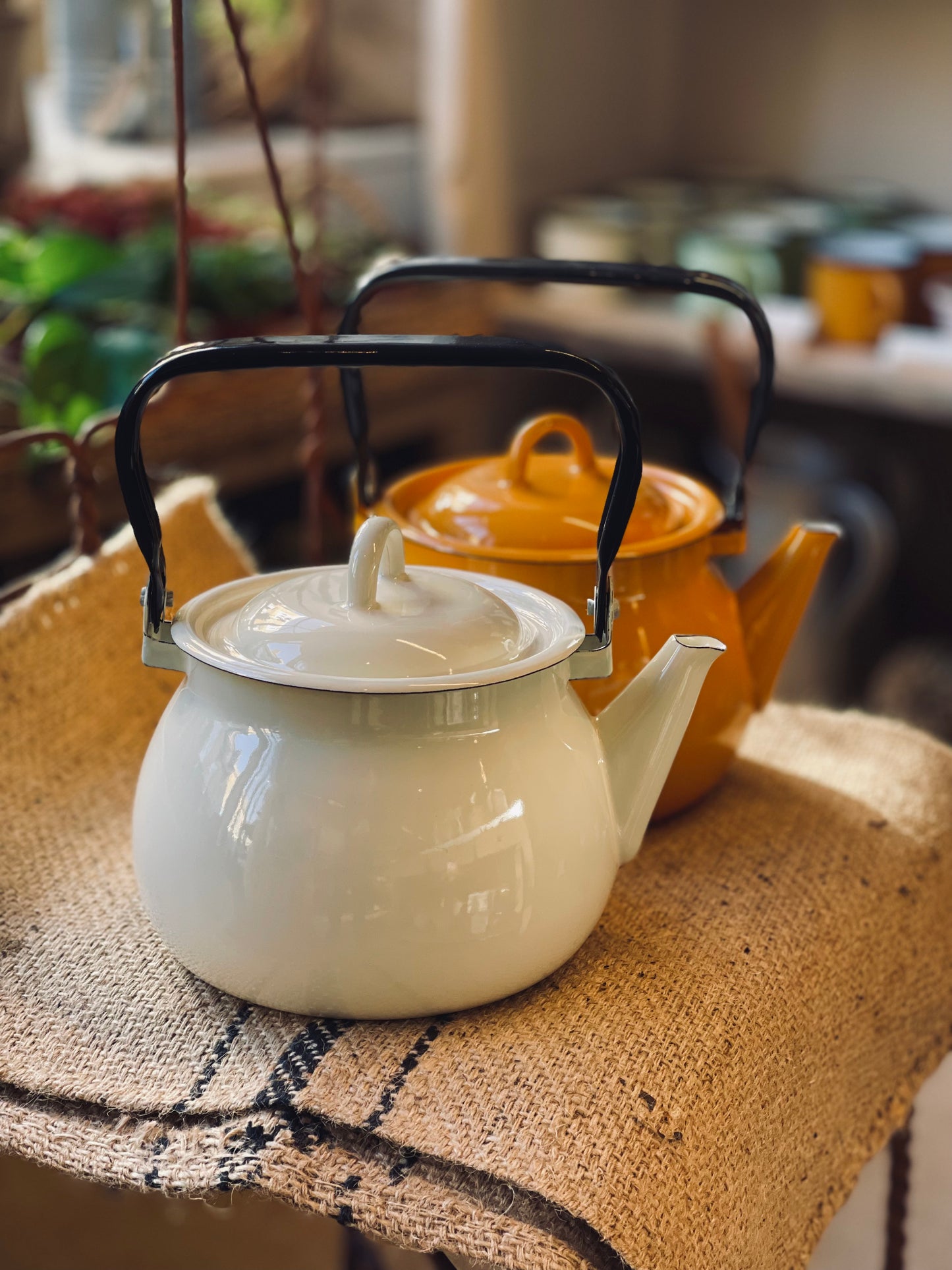 Two kettles, Ukrainian made with enamel, one white and one yellow with a black handle, on a textured surface.