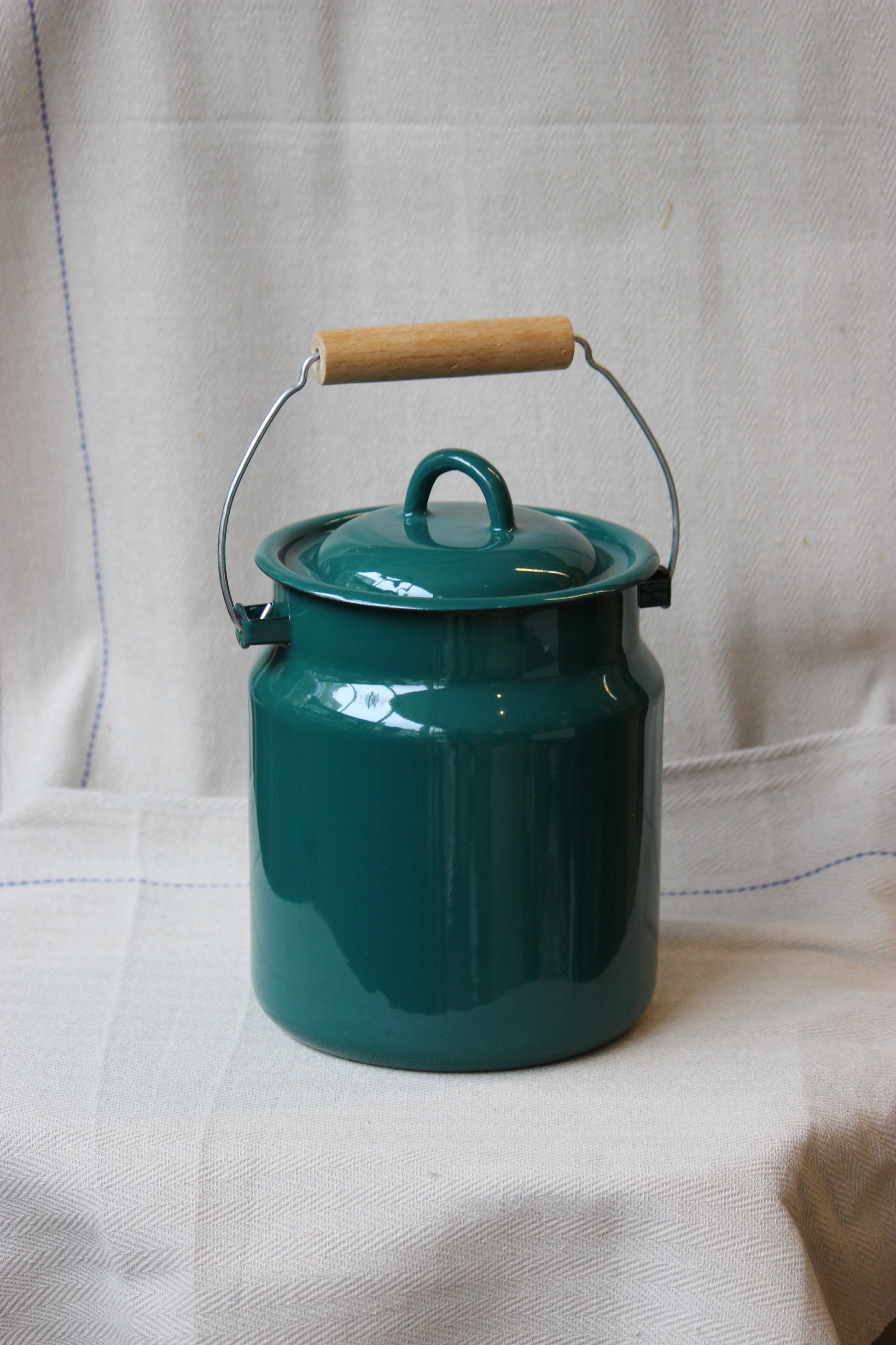 Teal enamelware storage jar made in Ukraine with wooden handle on a white fabric background