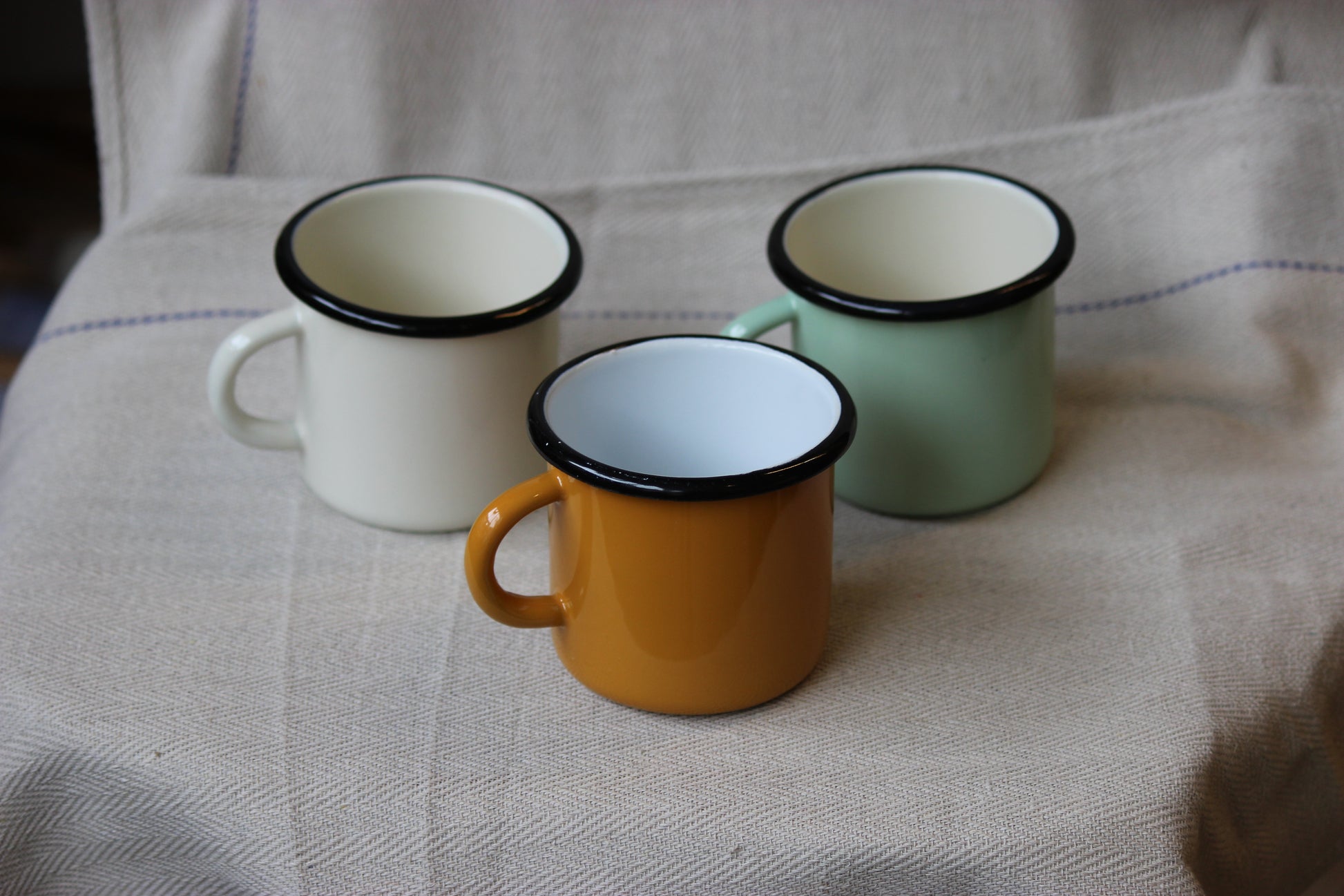 Three enamel mugs in white, green, and orange on a textured fabric surface.