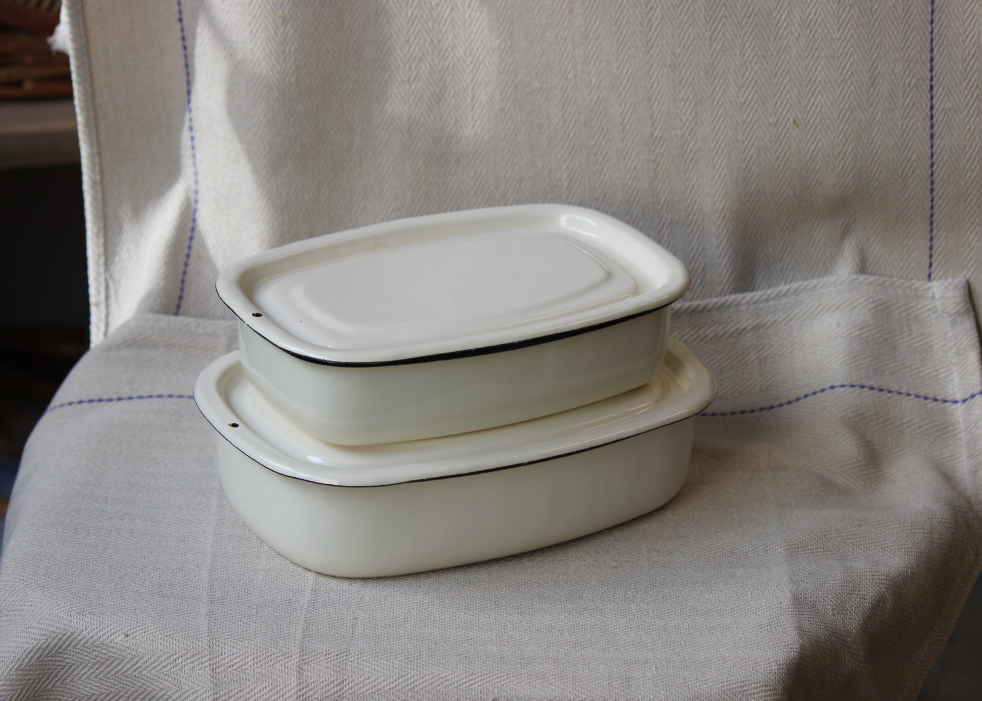 Two white enamel dishes with lids on a textured fabric surface