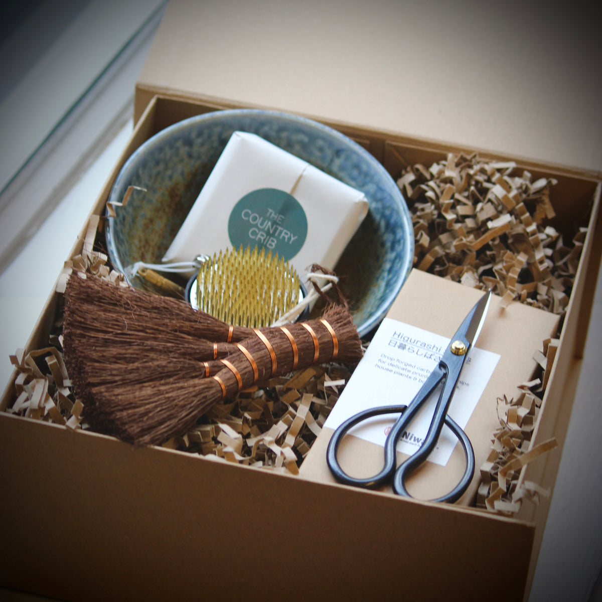 A curated gift box set containing a handmade brown broom, a blue speckled bowl, a gold-handled brush, a white soap bar labeled 'The Country Crib', and silver scissors, all arranged in brown paper shred inside a kraft box.