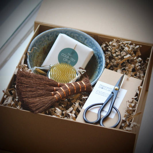 A curated gift box set containing a handmade brown broom, a blue speckled bowl, a gold-handled brush, a white soap bar labeled 'The Country Crib', and silver scissors, all arranged in brown paper shred inside a kraft box.
