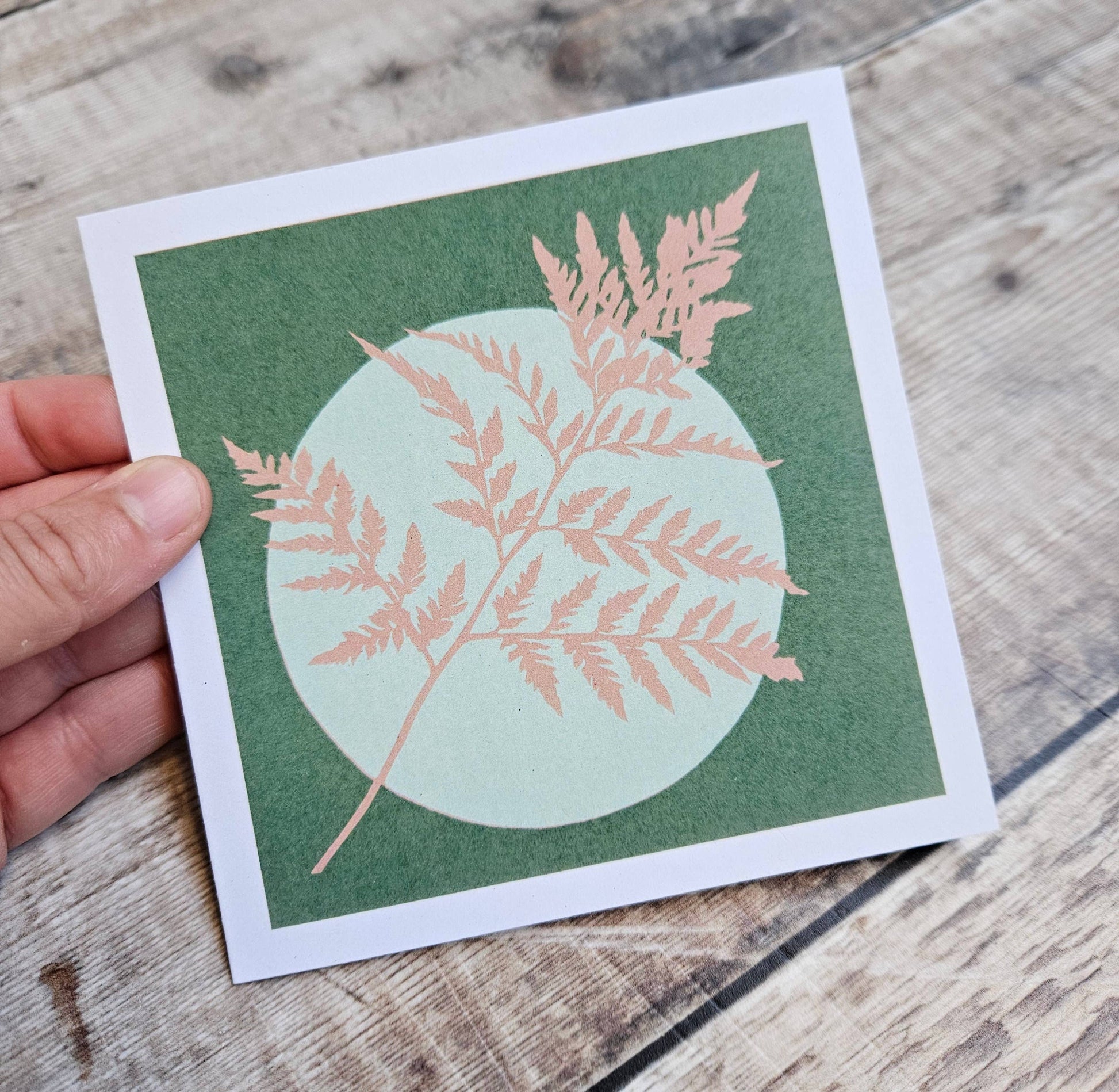 Card with a pink fern design on a green background held by a hand on a wooden surface