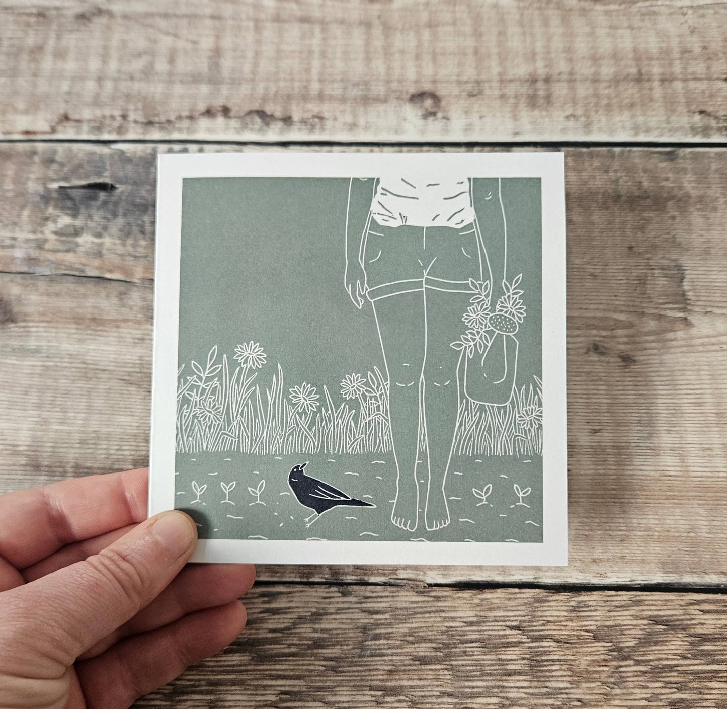 Hand holding a card with a design of a person holding flowers and a bird on a wooden surface