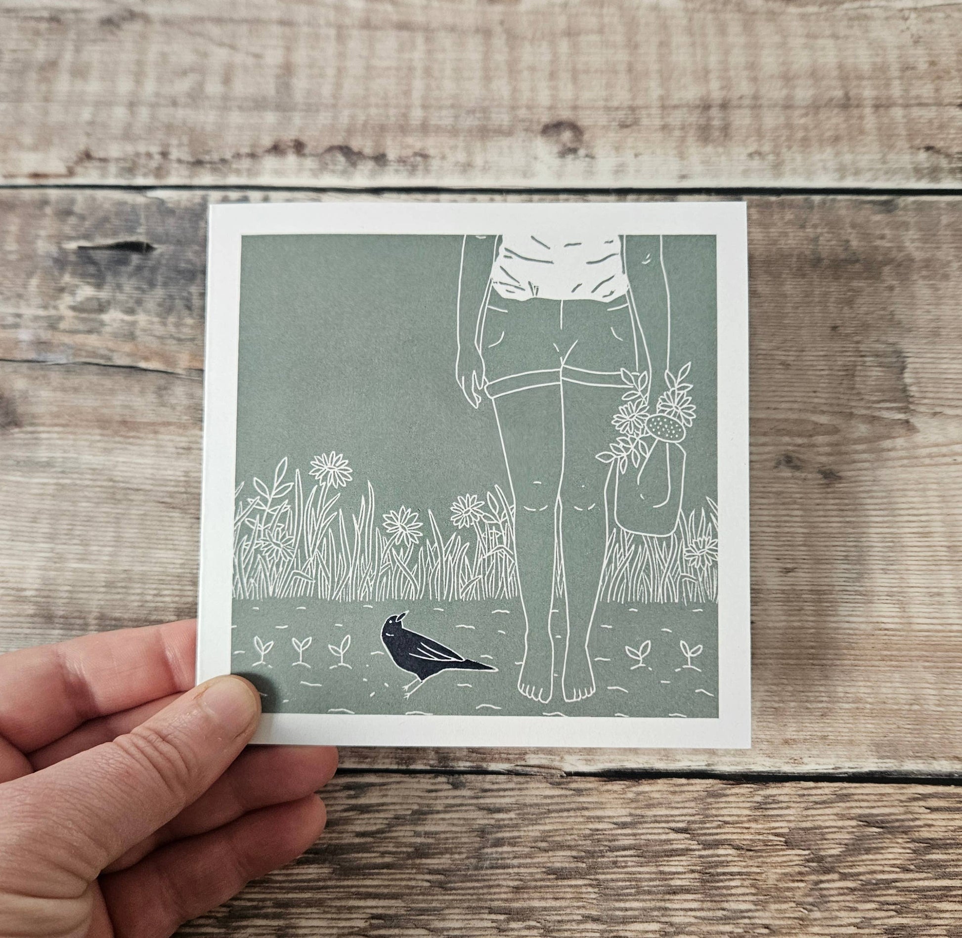 Hand holding a card with a design of a person holding flowers and a bird on a wooden surface