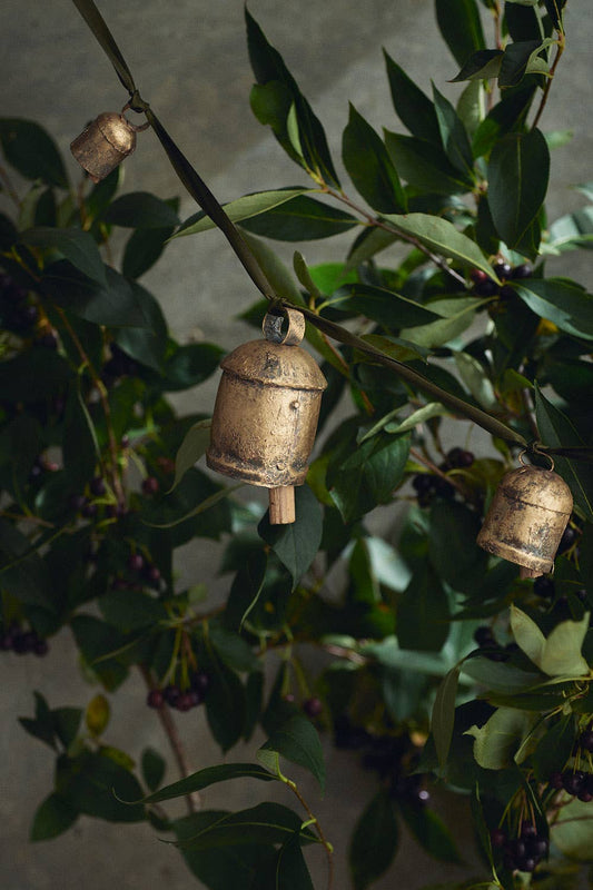 A bronze colored metal bell ornament hanging among green leaves, likely used as a Christmas decoration.