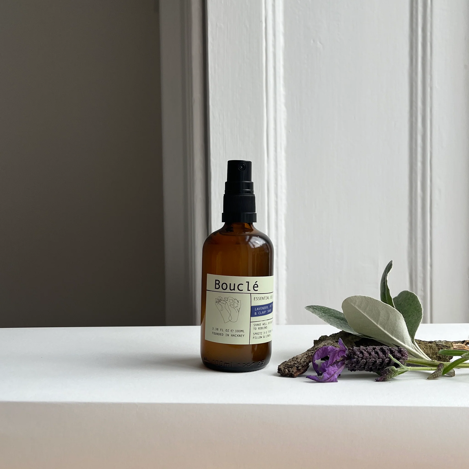 Bottle of Boucle on a white surface with lavender and eucalyptus leaves.