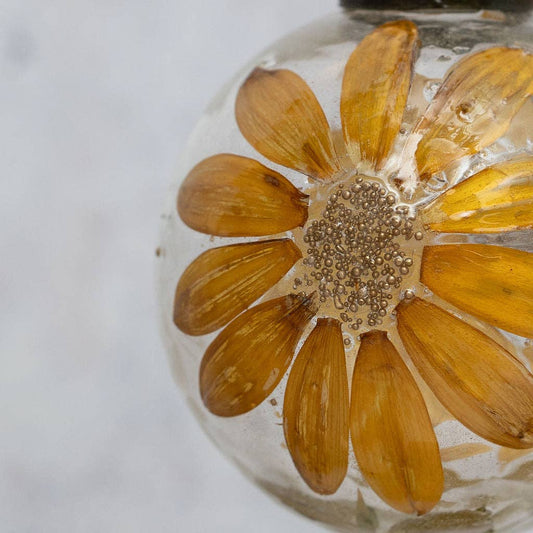 A close-up of a clear glass decorative bulb containing a pressed dried yellow-orange flower with a metallic silver center, set against a soft light gray background.