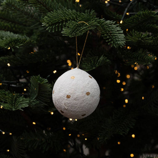 A cotton paper mache white textured spherical Christmas ornament with small gold polka dots hangs from a gold string on a dark green Christmas tree with warm glowing lights.