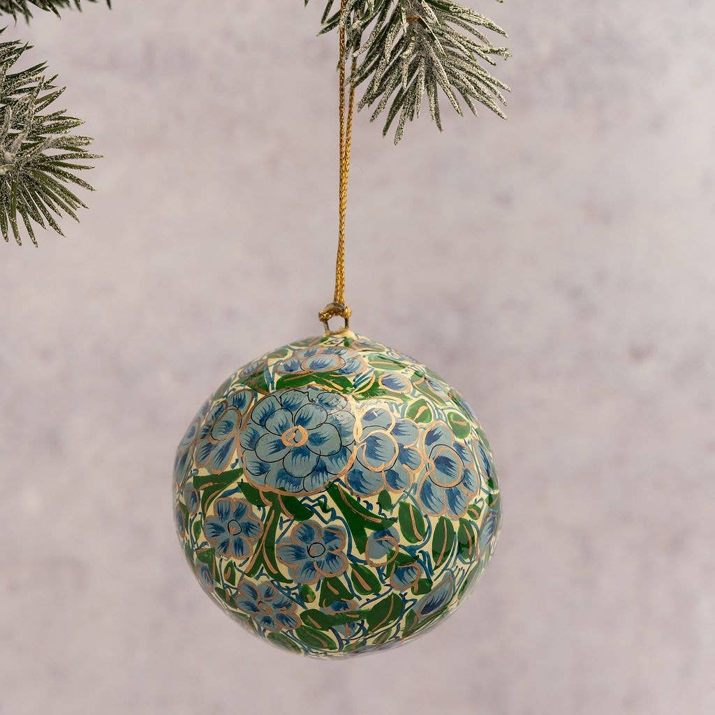 A round glass Christmas ornament decorated with a hand-painted blue and green floral pattern, hanging from a gold string and suspended from a pine branch against a soft, light gray background.