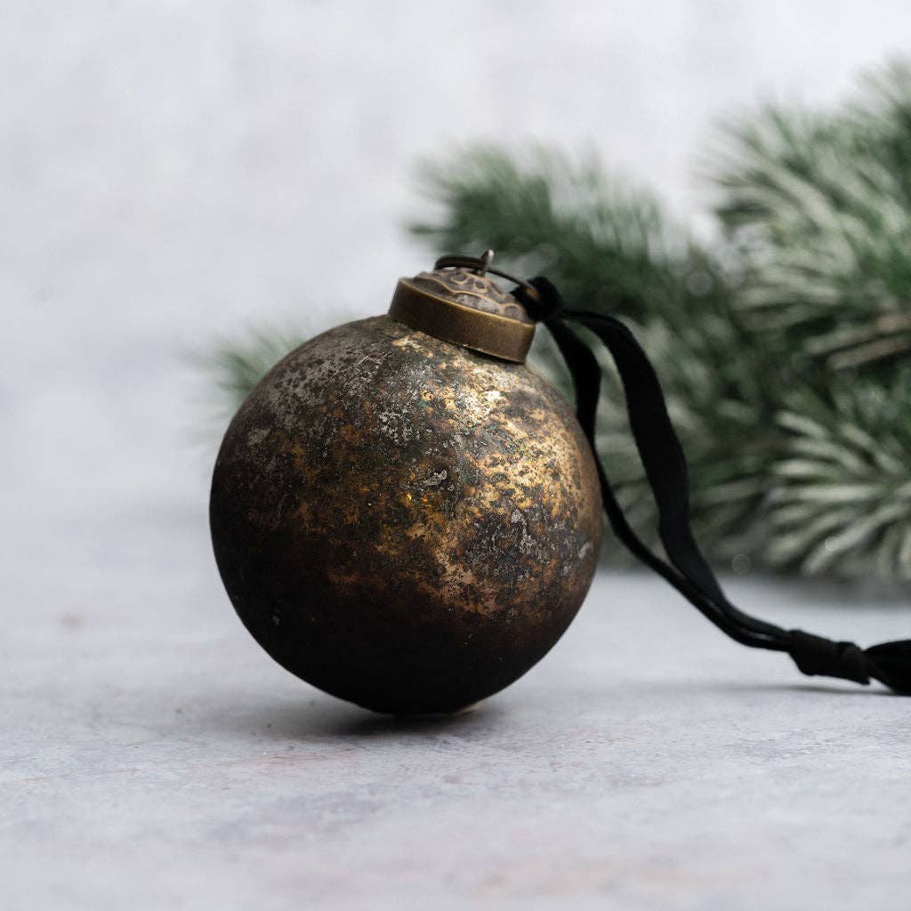 A round metallic Christmas ornament with a distressed gold and black finish, hanging from a black ribbon, set against a soft gray background with blurred green pine branches.