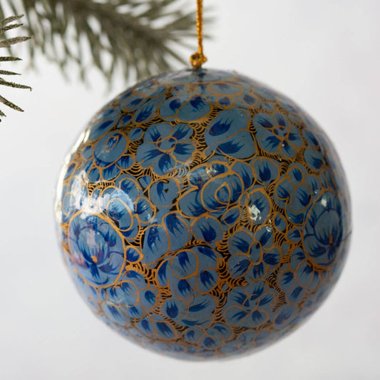 A glossy blue paper mache Christmas ornament decorated with intricate gold floral patterns, hanging from a gold string against a white background with a pine branch visible in the upper left.