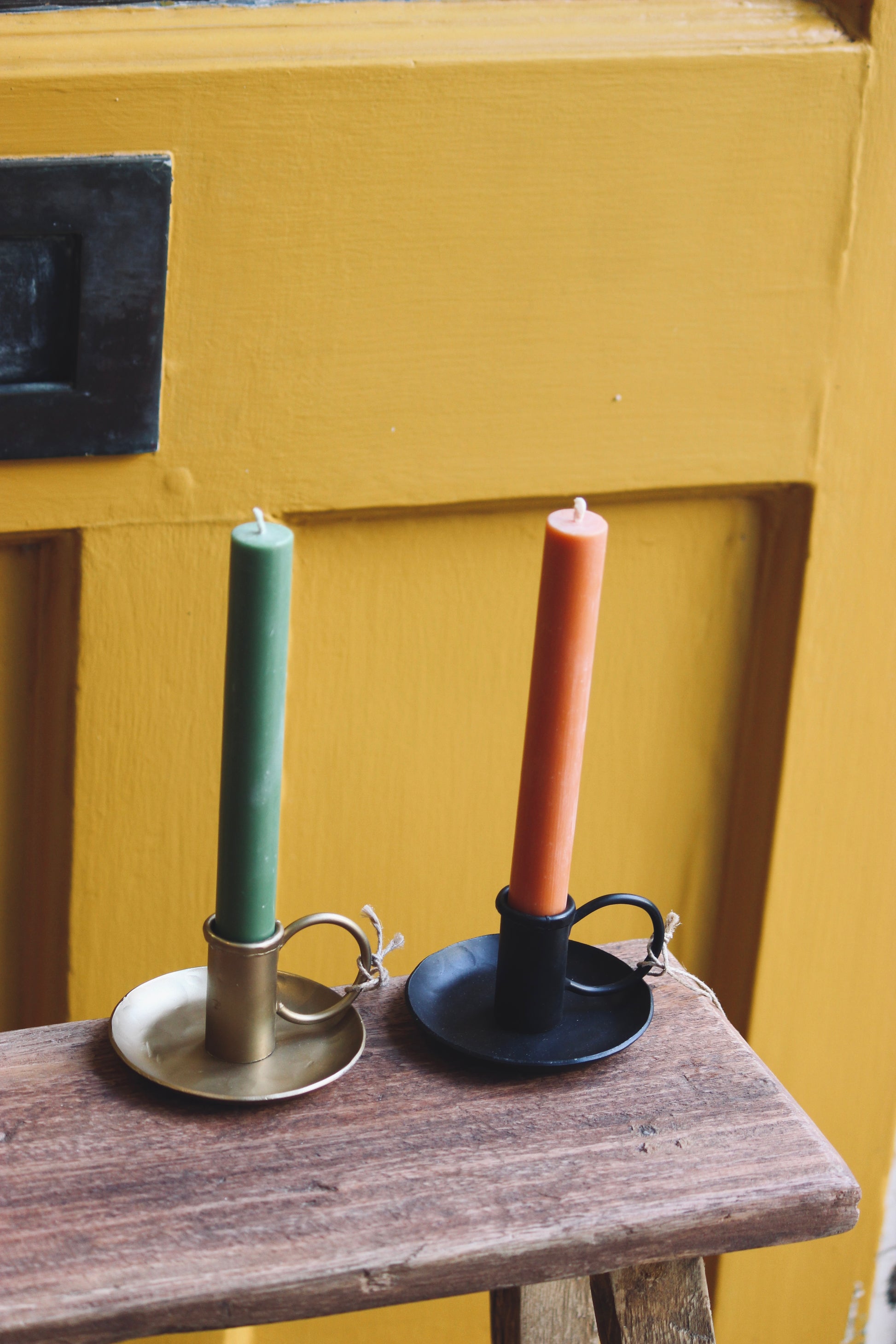 Two candles in nkuku iron candle holders on a wooden surface with a yellow door background.
