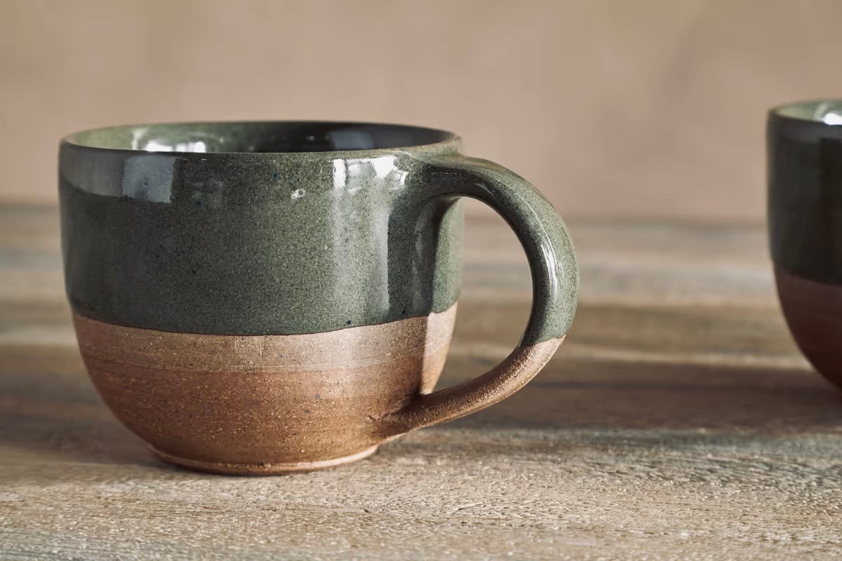 A nkuku mali ceramic mug with a two-tone glaze, featuring a dark olive green upper half and a natural brown clay lower half, set on a rustic wooden surface.