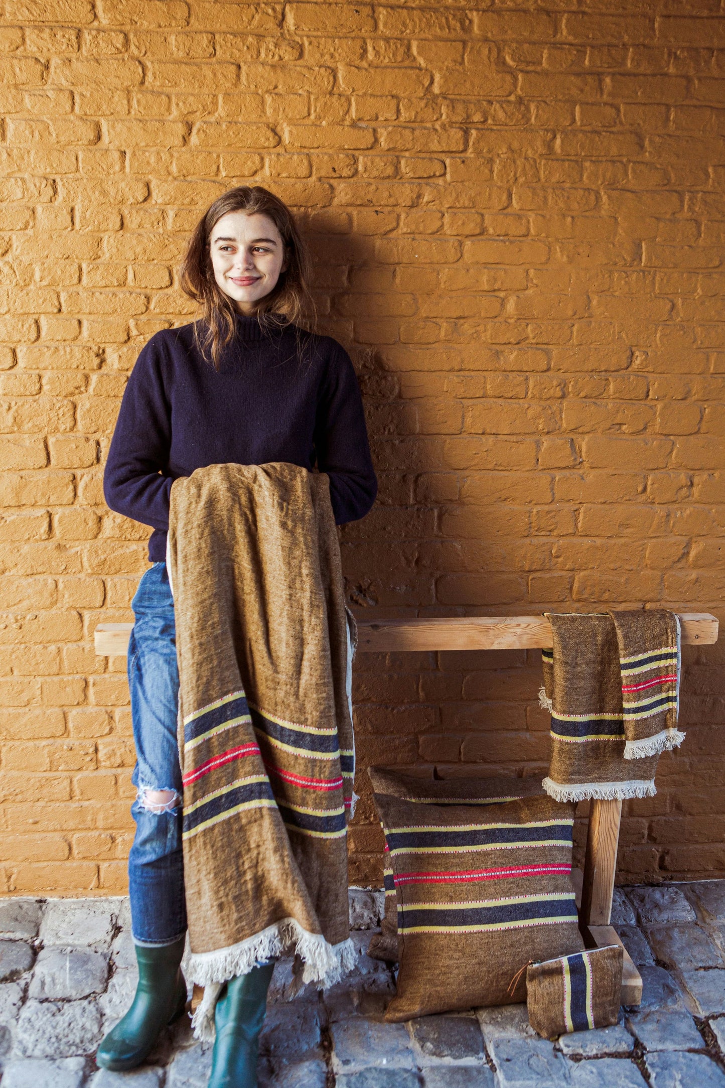 Woman holding a Libeco woven blanket against a brick wall with similar items on a table.