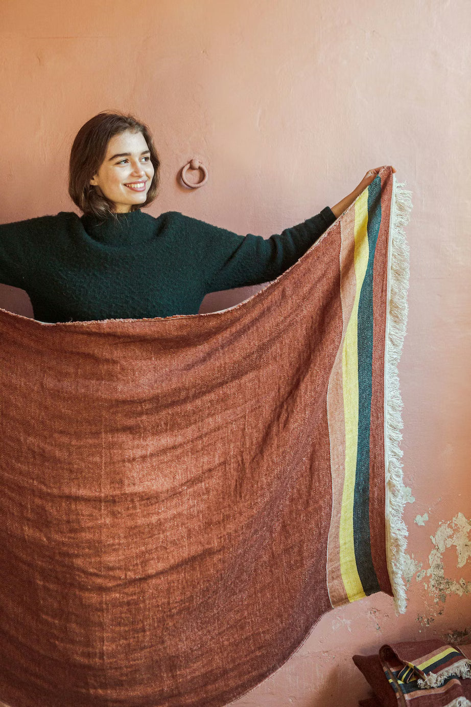 Woman holding a large brown fabricLibeco - The Belgian Towel Fouta 100% Linen  with colorful stripes against a pink wall.